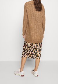 Donna che indossa un maglione di lana marrone, una gonna midi con stampa leopardata e sneakers bianche, in piedi di fronte a una parete bianca semplice.