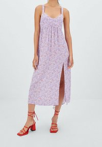 Femme portant une robe sans manches à fleurs lilas avec une fente haute sur la cuisse et des sandales à talons blocs rouges à lanières, se tenant devant un fond uni.