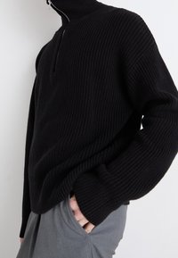 Schwarzer Strickpullover mit hohem Kragen und Reißverschluss, der ein strukturiertes Rippmuster und eine Übergröße aufweist. Graue Hose an der Unterseite sichtbar.