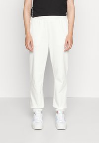 Frau in weißen Jogginghosen mit elastischen Bündchen und weißen Adidas-Sneakern mit hellblauen Streifen, stehend vor einem schlichten Hintergrund.