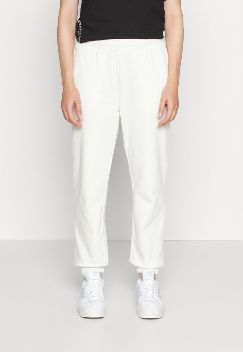 Frau in weißen Jogginghosen mit elastischen Bündchen und weißen Adidas-Sneakern mit hellblauen Streifen, stehend vor einem schlichten Hintergrund.