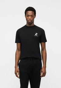 Άνδρας που φοράει μαύρο κοντομάνικο μπλουζάκι με λευκό λογότυπο φοίνικα και μαύρο παντελόνι με κορδόνι, στέκεται μπροστά σε απλό ανοιχτό φόντο.