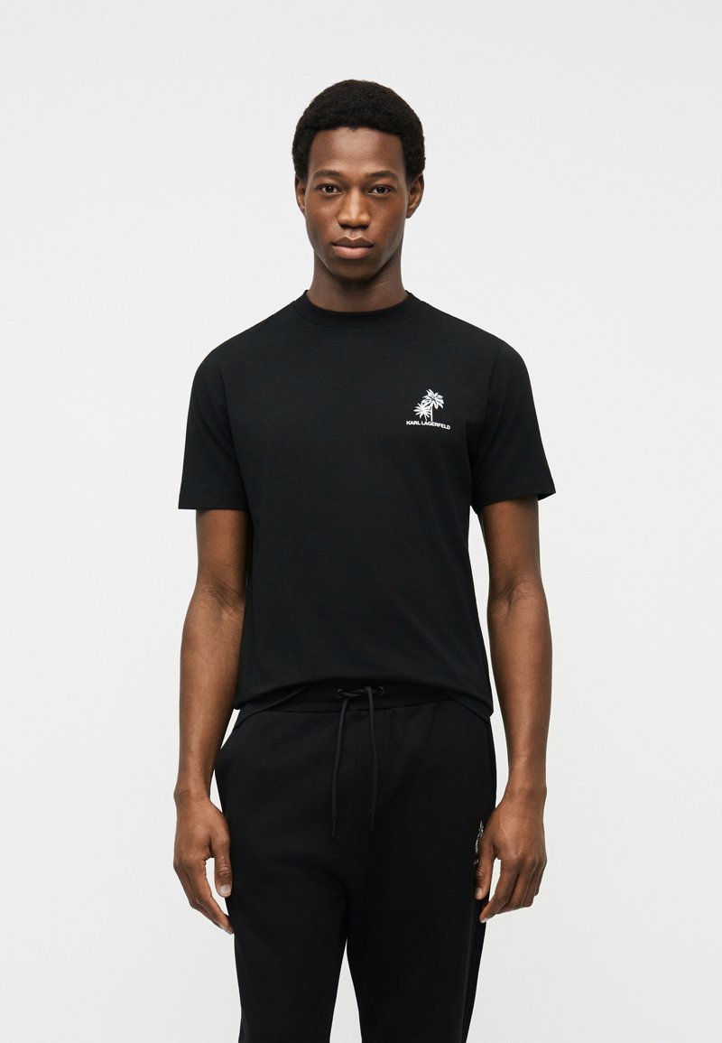 Άνδρας που φοράει μαύρο κοντομάνικο μπλουζάκι με λευκό λογότυπο φοίνικα και μαύρο παντελόνι με κορδόνι, στέκεται μπροστά σε απλό ανοιχτό φόντο.