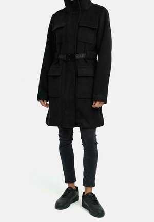 Person iført sort frakke med bælte og fire forlommer, sorte tætsiddende bukser og sorte sneakers, stående mod en ensfarvet lys baggrund.