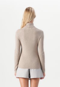 Kvinde med langt blont hår iført en beige ribbet sweater med høj hals og lysegrå shorts, stående med ryggen mod kameraet.