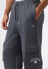 Mann trägt dunkelgraue Champion Cargo-Sweatpants mit Logo-Patch und Kordelzugbund, Hand in der rechten Tasche.