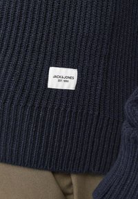 Maglione lavorato a maglia di colore blu scuro con texture a coste; presenta un'etichetta bianca con la scritta "JACK & JONES EST. 1990" attaccata all'orlo.