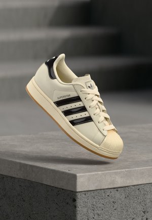 Zapatilla Adidas Superstar color crema con rayas negras y lengüeta en el talón, puntera de concha en crema y suela de goma color caramelo, mostrada en el aire sobre una superficie de concreto.
