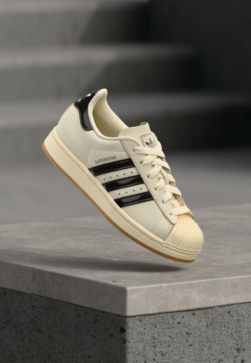 Baskets Adidas Superstar blanc cassé avec des bandes noires et un renfort talon noir, embout coquille crème et semelle en caoutchouc gomme, montrées en l'air au-dessus d'une surface en béton.