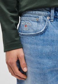 Jasnoniebieskie dżinsy z denimu mają naszywkę na tylnej kieszeni z napisem „Pepe Jeans London” i flagą Wielkiej Brytanii. Ręka umieszczona na talii.