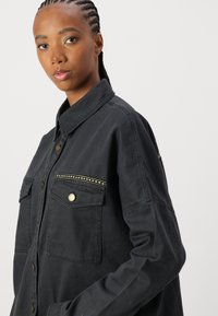 Giacca in denim nera con due tasche sul petto, bottoni in tonalità oro e cuciture decorative lungo la yoke delle tasche. Vestibilità leggermente oversize.