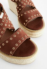 Sandali con zeppa in suede marrone, caratterizzati da cinturini incrociati e borchie dorate. La piattaforma in juta testurizzata aggiunge contrasto e stile.