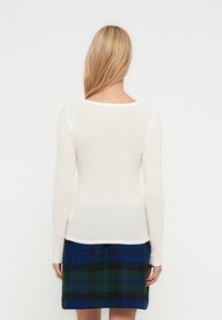 Μπλούζα με μακριά μανίκια σε εκρού χρώμα, με μαλακή, ελαφριά υφή. Συνδυασμένη με καρό φούστα σε μπλε και πράσινες αποχρώσεις, με ίσια γραμμή και τελείωμα.