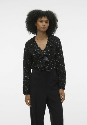 Blusa nera con pois bianchi, scollatura arricciata e maniche lunghe. Abbinata a pantaloni neri a vita alta. Texture del tessuto liscia.