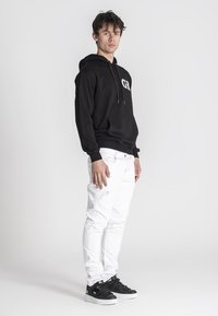 Čierna mikina s kapucňou a prednou vreckom, skombinovaná s bielymi obtiahnutými džínsami a čiernymi teniskami s bielymi podošvami. Minimalistický dizajn.