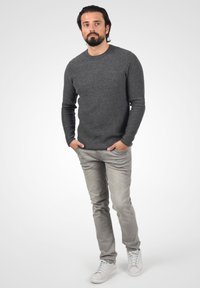Suéter gris de punto, jeans gris claros y zapatillas blancas. Hombre de pie con las manos en los bolsillos, posando sobre un fondo gris claro.