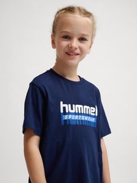 Uśmiechnięte dziecko z blond włosami w warkoczach, ubrane w granatowy T-shirt „hummel SPORTSWEAR”, stojące na jednolitym jasnym tle.