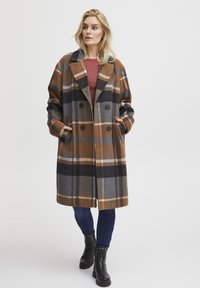 Double-breasted plaid coat in brown, gray, and navy, featuring large buttons, a notched collar, and side pockets, worn over a maroon top.