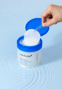 Genomskinlig rund behållare med blått lock, som innehåller vita texturerade bomullspads. En hand lyfter en pad från förpackningen över en vågig blå yta.