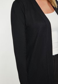 Cardigan nero con una texture liscia, apertura frontale e design a V. Indossato sopra un top bianco, mette in evidenza il contrasto di colori.