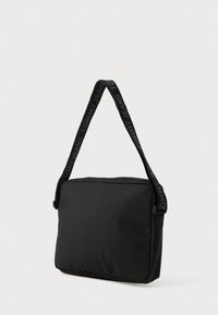 Sort rektangulær skuldertaske lavet af stof, med en tekstureret rem hvor "TOMMY JEANS" er trykt langs længden, samt en lynlåslukning.