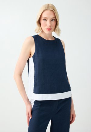 Donna bionda che indossa un top blu navy senza maniche con pannelli laterali bianchi e pantaloni blu navy abbinati, in piedi su uno sfondo chiaro.
