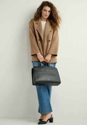 Młoda kobieta w beżowym płaszczu dwurzędowym, niebieskich dżinsach, czarnych butach na klockowym obcasie, trzymająca dużą czarną skórzaną torbę na tle jednolitego tła.