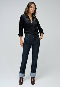 Camisa negra de satén con botones y mangas dobladas, combinada con jeans de mezclilla oscuros con dobladillos y botas de tacón marrones.