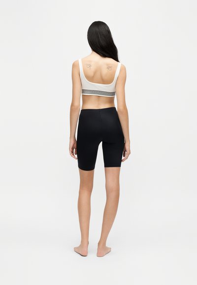 Femme aux longs cheveux foncés, debout pieds nus, de dos, portant un crop top blanc et un short noir taille haute, montrant des tatouages symétriques d’yeux sur le haut du dos.