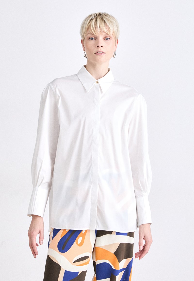 Camisa blanca con cuello abotonado, mangas largas abullonadas y textura suave; combinada con pantalones de patrones coloridos que presentan diseños en azul y naranja.
