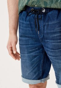 Hombre con pantalones cortos de mezclilla enrollados y cinturilla de cordón negra, llevando una camisa verde con un patrón metida por dentro.