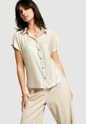 Femme portant un chemisier brillant à manches courtes et boutonné ainsi qu’un pantalon ample beige clair, posant avec la main dans la poche sur un fond uni.