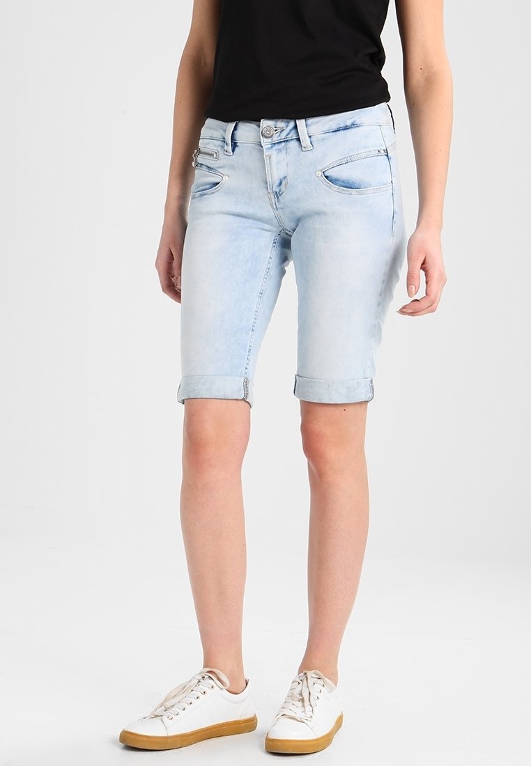Kvinna som bär ljusblå, uppvikta denimshorts, vita sneakers med gummisula och en svart ärmlös topp mot en enfärgad bakgrund.