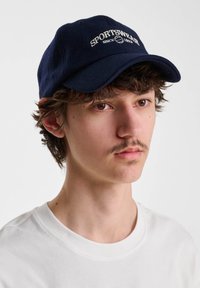 Ung man med lockigt hår som bär en marinblå keps med texten "SPORTSWEAR sedan 1929" och en enkel vit t-shirt på en vit bakgrund.