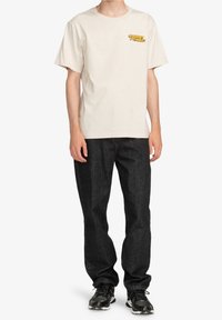 Světle béžové bavlněné tričko se žlutým grafickým logem na levé straně hrudi, v kombinaci s černými džíny a černými teniskami.