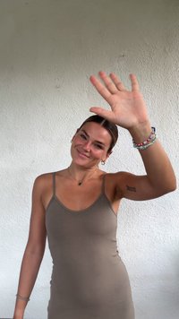 Femeie cu păr brunet de lungime medie, purtând un maiou bej, care saluta cu mâna dreaptă. La încheietura stângă are mai multe brățări colorate.