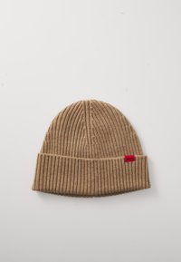 Tkanja beanie v svetlo rjavi barvi z rebrasto teksturo, zložljivim robom in majhno rdečo nalepko. Mehka tkanina, zaobljena oblika, primerna za hladno vreme.