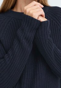 Pull en maille navy avec un motif texturé à côtes ; poignets larges et col rond. Le matériau semble doux et épais.