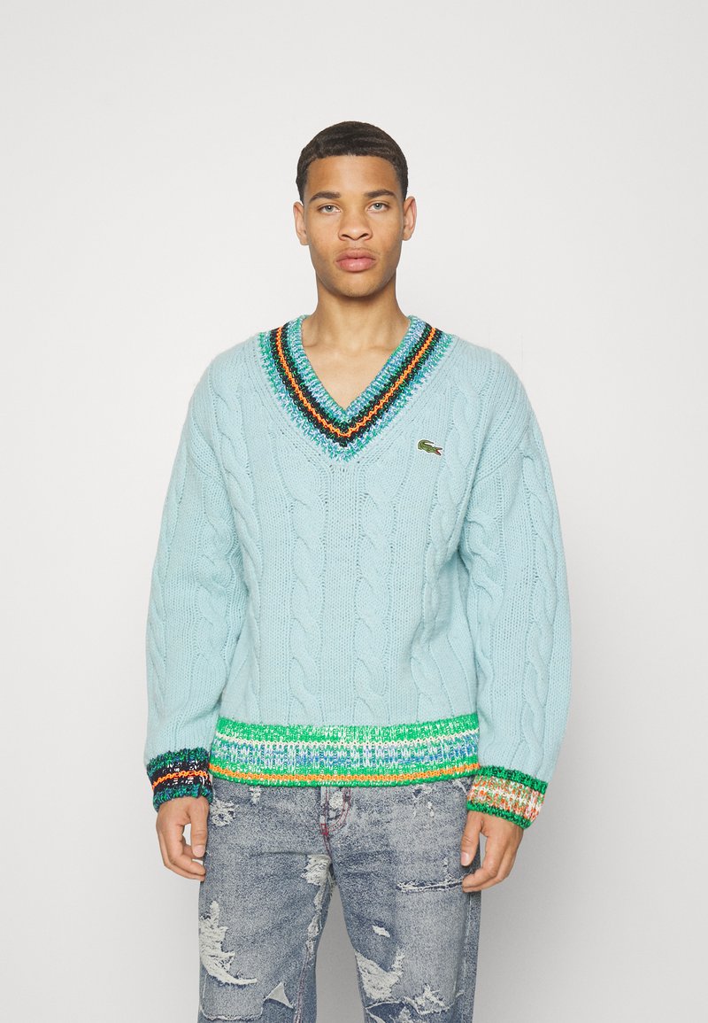 Maglione azzurro chiaro a trecce con scollatura a V, caratterizzato da accenti striati multicolori sul colletto e sull'orlo. Indossato con jeans in denim strappati.