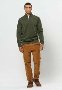Homme debout portant un pull vert olive à demi-fermeture éclair, un pantalon en velours côtelé marron et des bottes marron à lacets sur un fond clair uni.