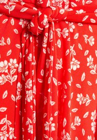 Czerwona tkanina z białym kwiatowym wzorem, z detalem związania w talii. Miękka tekstura, lekki materiał i swobodny krój.