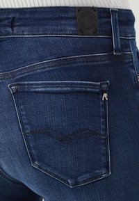 Jeans de mezclilla azul oscuro con un bolsillo trasero que presenta un patrón de puntada en zigzag y una etiqueta negra cerca de la cintura para la identificación de la marca.