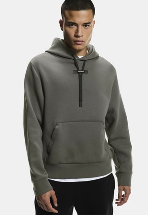 Jonge man die een grijze hoodie draagt met zwarte trekkoorden en een voorzak, staand tegen een effen achtergrond.