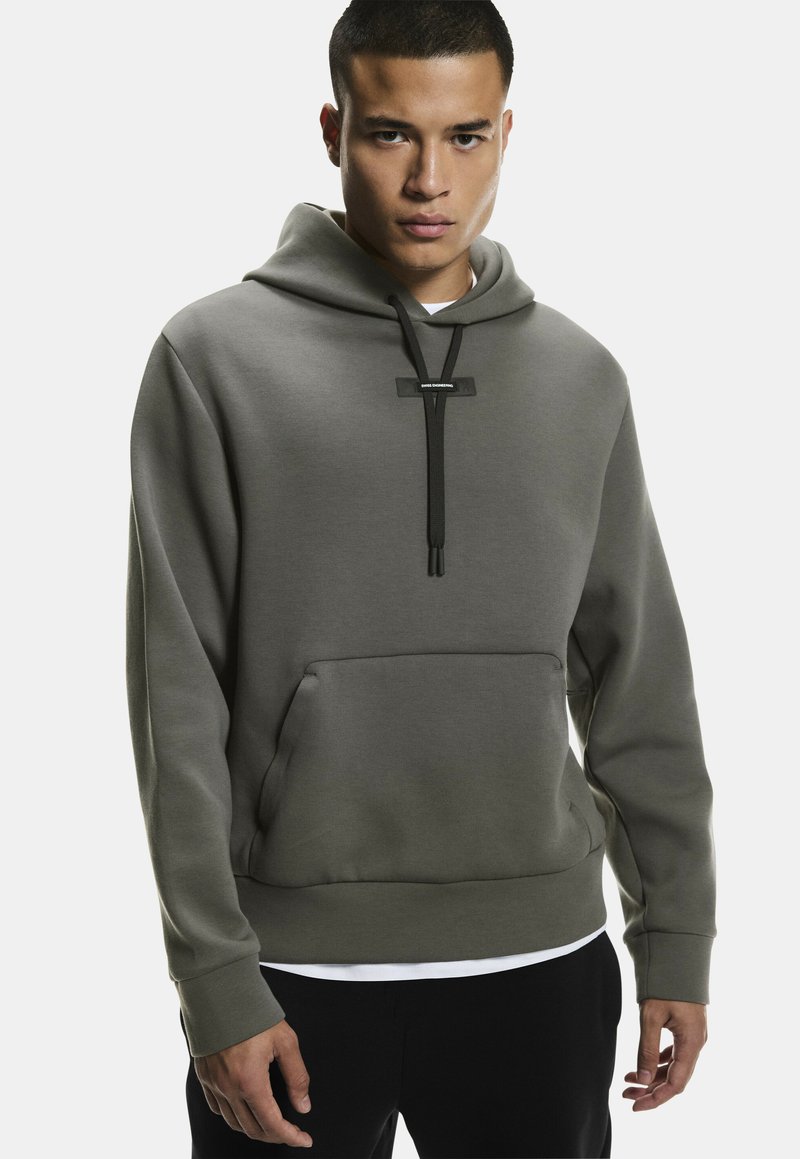 Jeune homme portant un sweat à capuche gris avec des cordons noirs et une poche kangourou à l'avant, debout devant un fond uni.