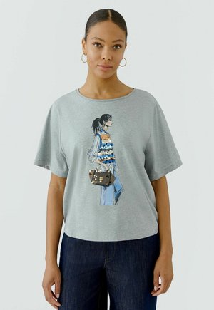 Vrouw met donker haar naar achteren gebonden, draagt gouden creolen, een grijs T-shirt met een kleurrijke paillettenafbeelding en donkerblauwe spijkerbroek tegen een effen achtergrond.