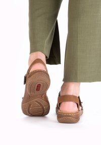 Brown sandals made of textured material, featuring back strap closure and a patterned rubber sole, paired with olive green pants.