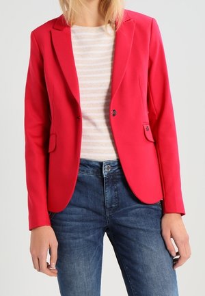 Donna che indossa una giacca rossa aderente sopra una camicia a righe bianche e beige con jeans di denim blu, in piedi contro uno sfondo chiaro.