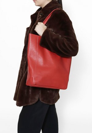 Red leather tote bag with a textured surface and sturdy straps, held by a person wearing a brown coat. Simple, rectangular shape.