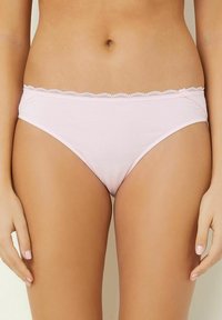 Donna che indossa lingerie di cotone leggero con pizzo rosa chiaro, in piedi con le mani rilassate lungo i fianchi su uno sfondo semplice.