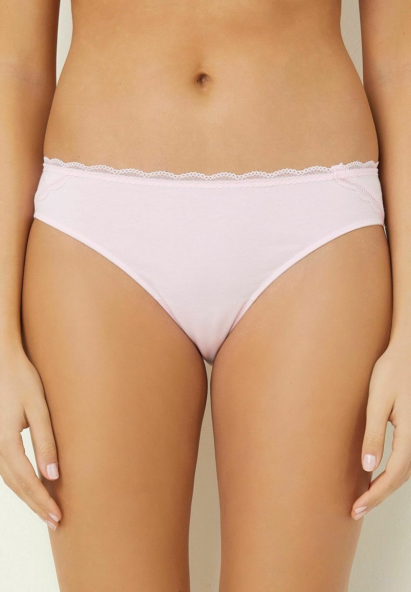 Donna che indossa lingerie di cotone leggero con pizzo rosa chiaro, in piedi con le mani rilassate lungo i fianchi su uno sfondo semplice.